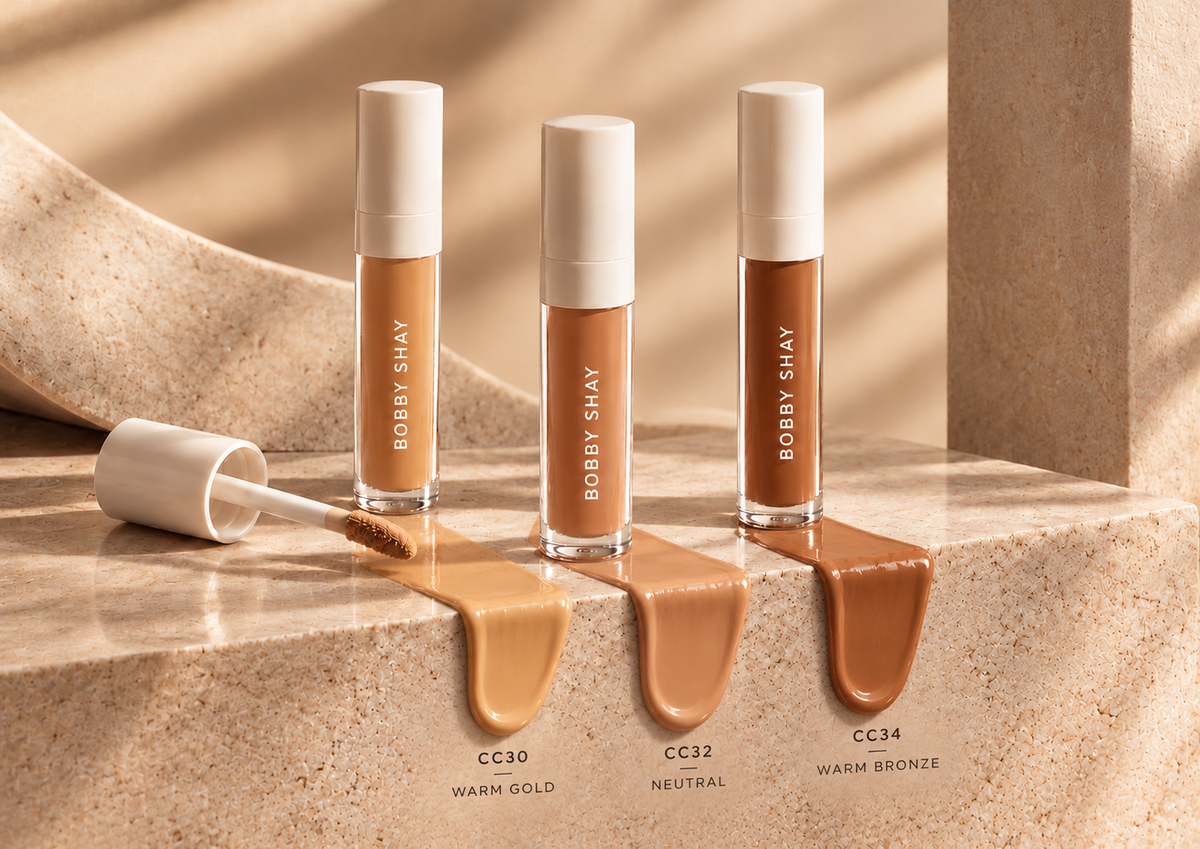 Three Bobby Shay Beauty liquid concealers in CC30 Warm Gold, CC32 Neutral and CC34 Warm Bronze, with white caps and formula swatches flowing over the edge of warm champagne stone