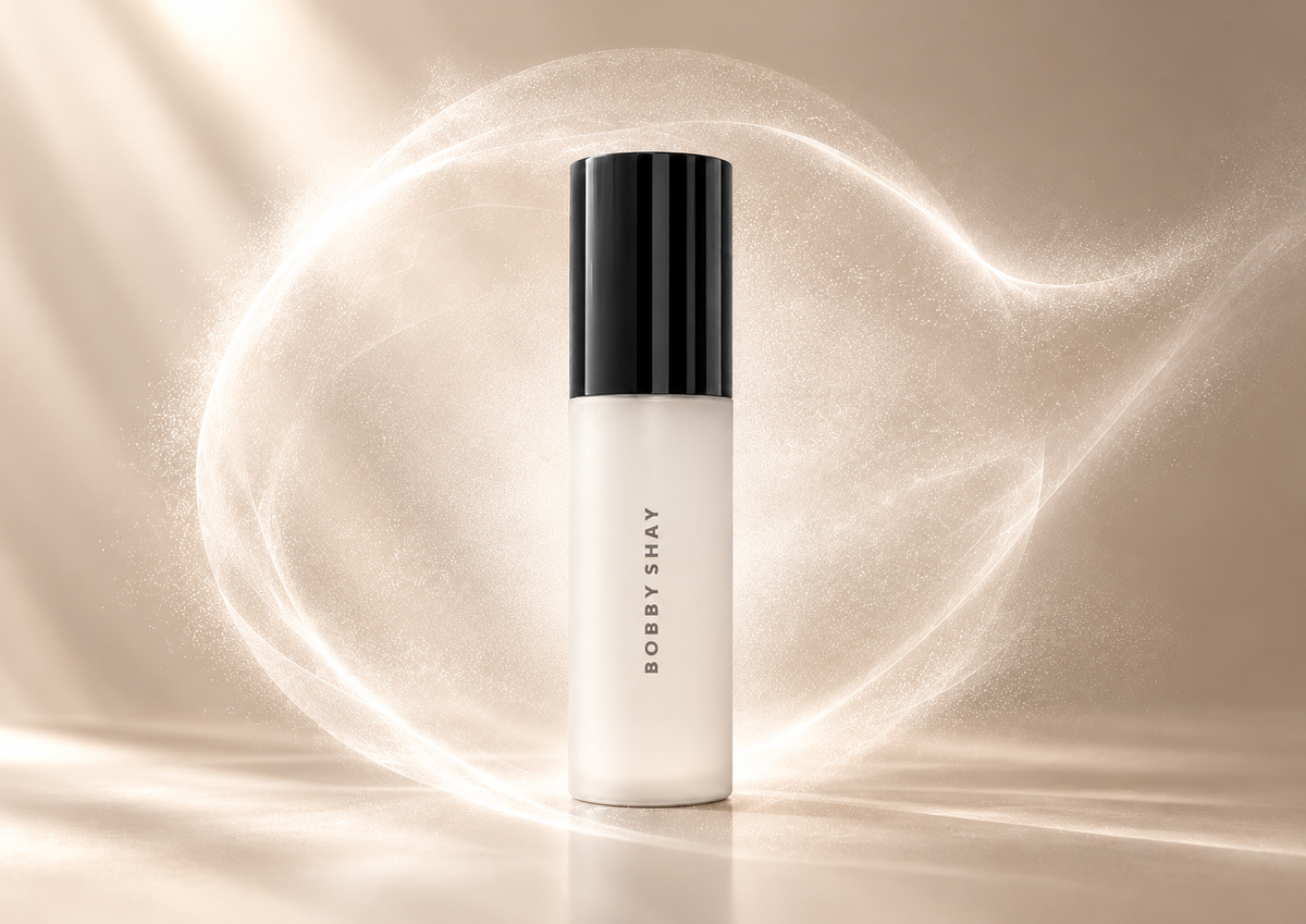 Bobby Shay Beauty Mist Me Baby setting spray in its frosted-white bottle and black cap, encircled by a delicate mist halo against a champagne background