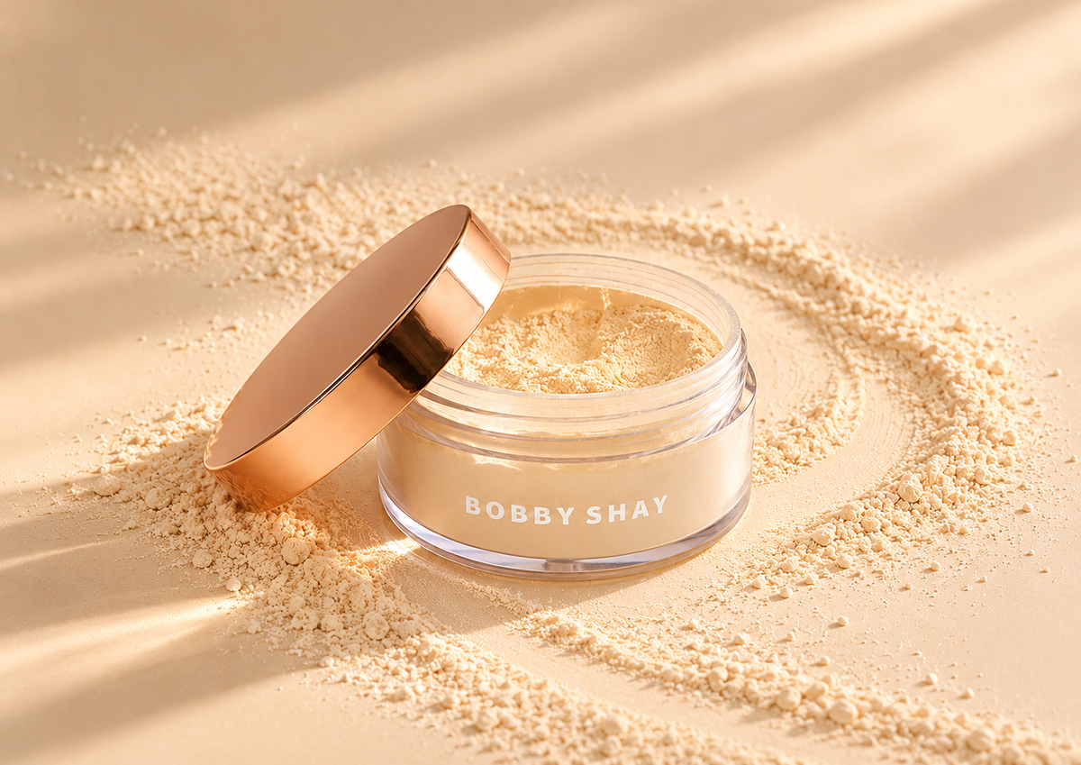 Bobby Shay Beauty Setting Veil loose powder in an open clear low-profile jar with rose-gold lid, surrounded by a soft, sweeping trail of powder on a champagne background